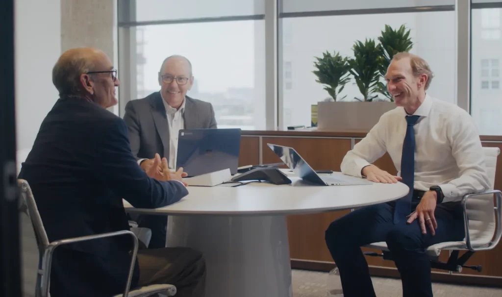 Three businessmen in professional attire having a meeting around a round table in a modern office with large windows
