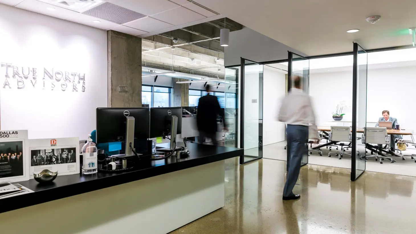 Modern office reception area with True North Advisors signage, front desk with monitors, and person walking toward glass-walled meeting room.