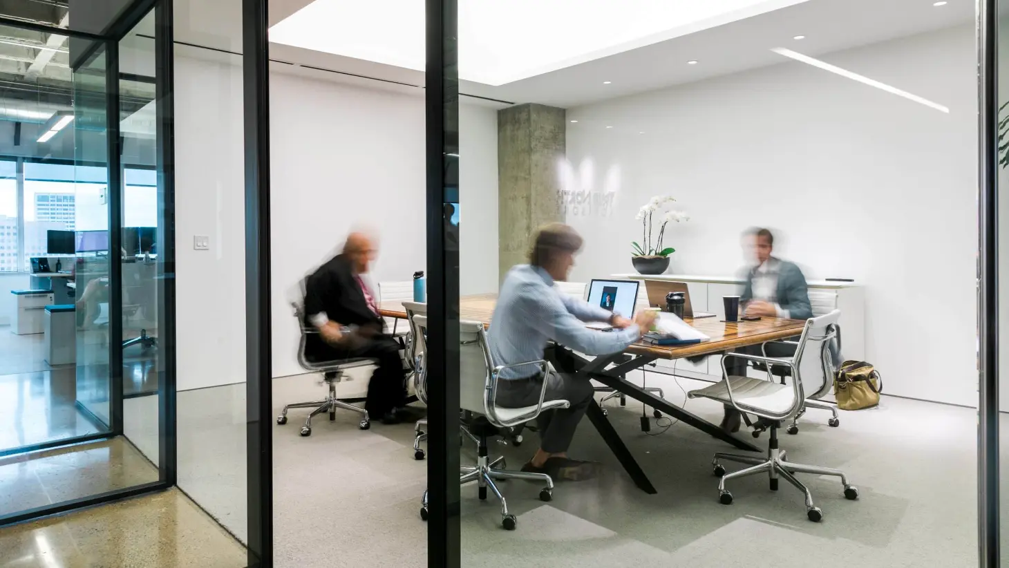 Three professionals meet in a modern glass-walled conference room at a wealth management office