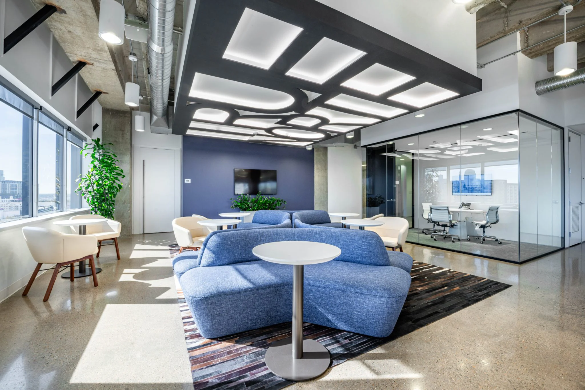 Modern office lounge with blue curved sofa, geometric ceiling lights, glass-walled conference room, and polished concrete floors.